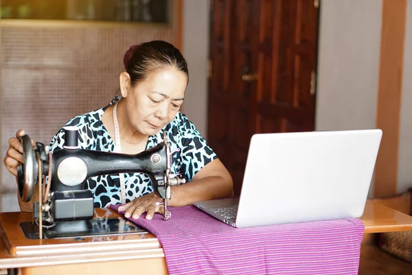 stock image Asian senior woman tailor is sewing cloth by using sewing machine, learn to design from laptop computer. Concept senior lifestyle spend free time to learn from online media      