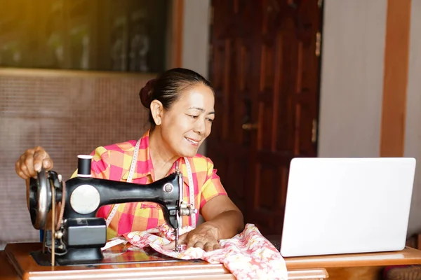 Asian senior woman tailor is sewing cloth by using sewing machine, learn to design from laptop computer. Concept senior lifestyle spend free time to learn from online media
