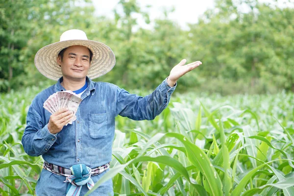 Happy Asian man farmer is at garden, wears hat, blue shirt, holds Thai banknote money, make hand gesture to present. Concept , Happy farmer to get profit, income, agriculture supporting money.