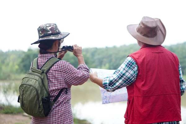 Back view of Asian woman and man travellers holds binocular and paper map to explore natural route for trekking beside the lake. Ecology study. Pastime activity, lifestyle. Explore environment