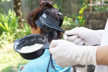 Closeup hands holds black brush to apply cream to dye hair of senior Asian woman, outdoor. Concept, Dye hair at home . Change hair color.      