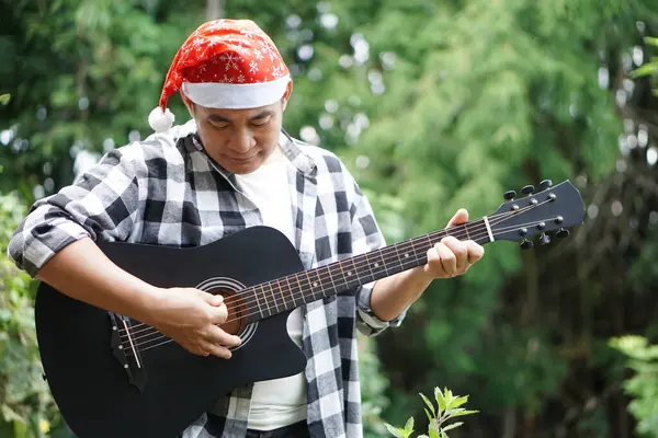Asian man is playing acoustic guitar in the park, outdoor nature background. Concept, love music, hobby, recreation activity. Relax time. Playing music for celebrate Christmas party.