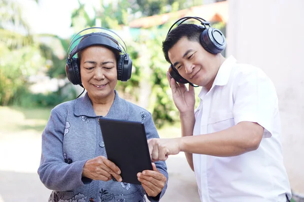Asian senior woman and Asian man wears headphones, holds smart tablet outdoor. Concept, son gives advice mom how to use online media from smart tablet. Learning new thing from internet.