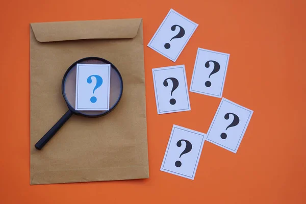 Brown envelope,magnifying glass and paper cards of question marks,orange background. Concept. Teaching aid. Education materials for doing activity or playing investigation games about find answers.