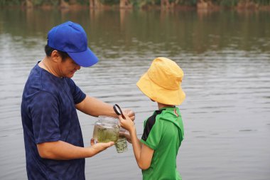 Asyalı baba ve oğul gölde ekolojiyi araştırıyor, baba cam su kavanozu tutuyor, oğul büyüteç tutuyor. Konsept, ekoloji dersi konusu doğa kaynaklı suyu inceler. Eğitim, yaşam deneyimi