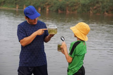 Asyalı baba ve oğul gölde ekolojiyi araştırıyor, baba cam su kavanozu tutuyor, oğul büyüteç tutuyor. Konsept, ekoloji dersi konusu doğa kaynaklı suyu inceler. Eğitim, yaşam deneyimi