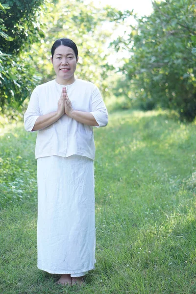 Asian woman Buddhist wears white costume, put palms together for praying, stands at tranquil forest. Full body image. Concept, culture of faith. Religious activity. Peace of mind management.