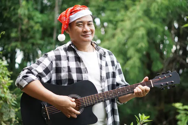 Asian man is playing acoustic guitar in the park, outdoor nature background. Concept, love music, hobby, recreation activity. Relax time. Playing music for celebrate Christmas party.