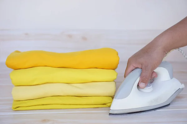 Stack of folded yellow clothes and hand holds electric iron on table. Concept, daily chore, household. ironing and folded clothes for neat and clean. Maintenance and keep garments.