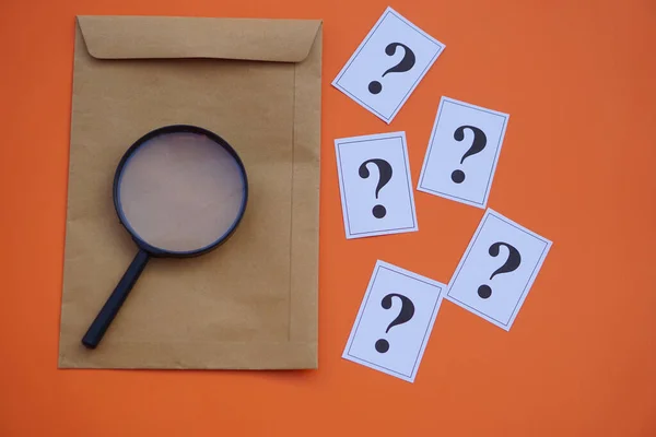 Brown envelope,magnifying glass and paper cards of question marks,orange background. Concept. Teaching aid. Education materials for doing activity or playing investigation games about find answers.