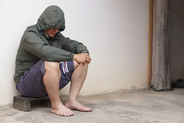 Depressed Asian man wear hoodie shirt, shorts, bare feet, put arms on knees, sits lean old wall on pavement. Concept, Life problems crisis from bad economy, poverty. Worried. Depression. Mental health
