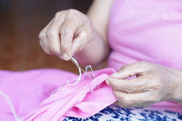 Close up senior woman hands uses needle and thread to repair clothes. Concept, hobby, free time activity. DIY craft. Fix and maintenance clothes by hand.  Sewing. Activity at home for elderly