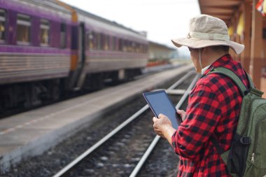 Asyalı yolcu tren istasyonunda, elinde akıllı tablet var. Konsept, Tayland 'da trenle seyahat, bilet almak, zaman çizelgesini kontrol etmek. Teknoloji taşımacılığı.          