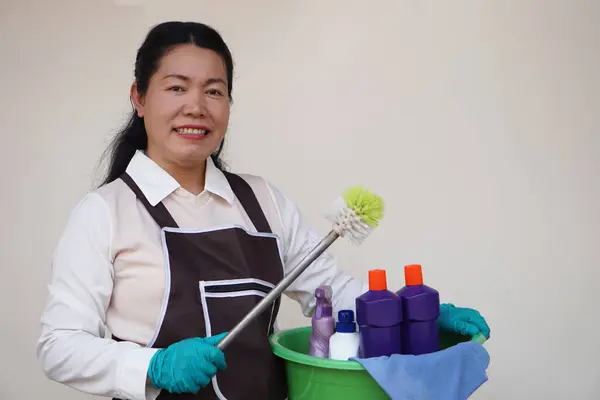 Asian woman housewife wears gloves, apron, holds bucket, bottles of cleaning liquid, alcohol spray, sanitary products and equipment for cleaning. Concept, Cleaning time for hygiene. Housekeeping.