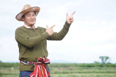 Yakışıklı Asyalı çiftçi tarım arazisinde duruyor, şapka takıyor, Tayland peştamalını beline takıyor, parmağını kaldırıyor, kendinden emin hissediyor. Kavram, tarım işgali. Taylandlı çiftçi.              