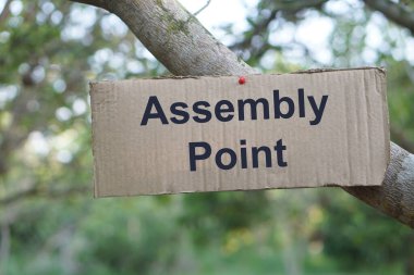 Üzerinde Assembly Point yazan kağıt kart tahtası, ağaca yapıştırılmış. İmza ve uyarı. Duyuru. Açık hava aktiviteleri için bilgi veriyor. Yürüyüş                