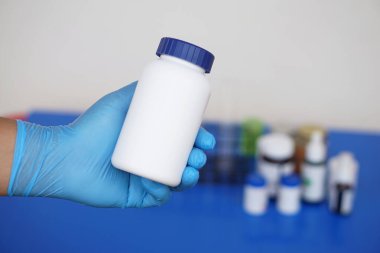 Kimyagerin eli koruyucu eldiven takar, beyaz bir şişe kimyasal ya da ilaç tutar. Kimyasal madde endüstrisi kavramı, eczane üretimi. Kimya laboratuvarında bilimsel deney.