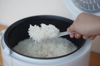 Eller elektrikli pilav pişirme makinesinden pirinç toplamak için plastik kaşık kullanır. Kavram, yemek pişirme, hizmet etmeden önce pirinç kalitesini kontrol etme. Yaşamak için günlük mutfak. Günlük hayatta elektrikli alet kullanın.