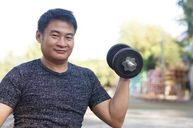  Yakışıklı Asyalı adam dışarıda egzersiz yapıyor, dambılı kaldırıyor. Konsept, vücut geliştirme egzersizi. Kaslarını güçlendir. Ağırlık kaldırma. Sağlıklı bir yaşam tarzı. Spor malzemeleri. Vücut çalışınca güçlenir..                                 