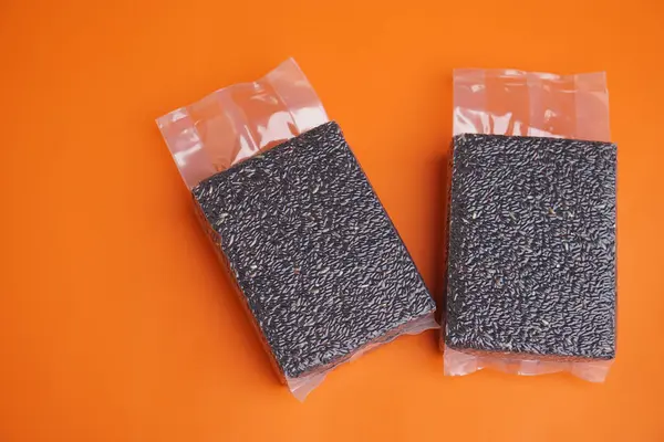 Turuncu arka planda, kapalı vakum torbasında iki paket 1kg siyah riceberry. Konsept, çevre dostu organik tahıl, besin açısından zengin, ihracat, sağlık piyasaları ve dünya çapında bilinçli tüketiciler için..                               