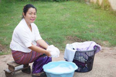  Geleneksel Tayland kıyafetleri içindeki Asyalı kadın mavi plastik bir leğende el yıkama, ahşap bir taburede sabun köpüğü ve çamaşır sepetiyle oturuyor. Tayland 'da kırsal yaşam.                                     