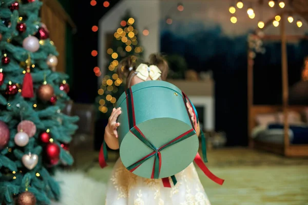 Child holding huge box with gift against backdrop of Christmas decorated at cozy home. Merry Christmas and Happy Holidays. Winter holidays, christmas and people concept. Girl carries Christmas present