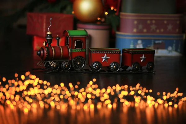 Red retro toy metal vintage soviet train with stars under Christmas tree on dark background near gifts, golden balls, bokeh lights garland on wooden rustic floor. New year, Holiday celebration concept