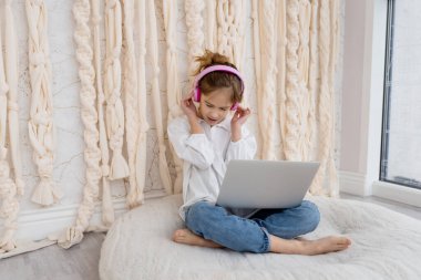 Caucasian funny child kid in headphones, sitting with computer, laptop at home. School girl, pupil having fun, using gadget, listening music, watching video class, playing video games. Young blogger.