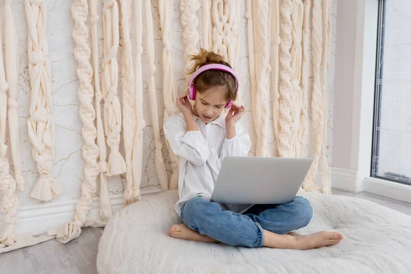 Caucasian funny child kid in headphones, sitting with computer, laptop at home. School girl, pupil having fun, using gadget, listening music, watching video class, playing video games. Young blogger.