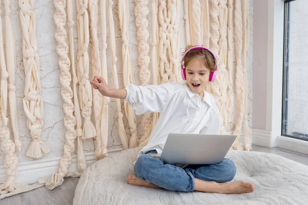 Caucasian funny child kid in headphones, sitting with computer, laptop at home. School girl, pupil having fun, using gadget, listening music, watching video class, playing video games. Young blogger.