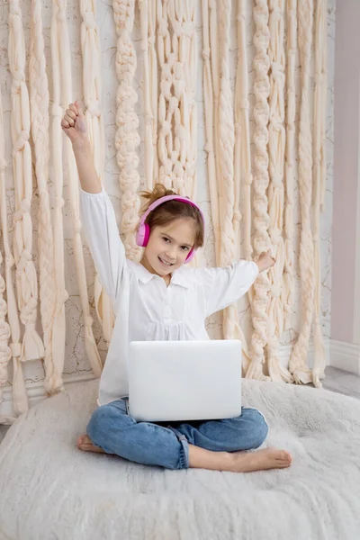 Caucasian funny child kid in headphones, sitting with computer, laptop at home. School girl, pupil having fun, using gadget, listening music, watching video class, playing video games. Young blogger.