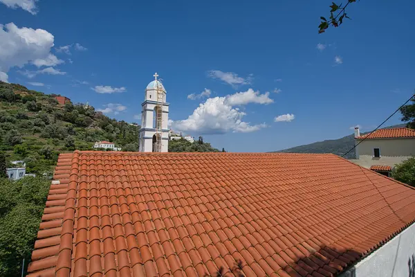 Andros Adası 'ndaki açık mavi gökyüzünün altında kırmızı kiremitli çatının arkasında haçlı beyaz çan kulesi.