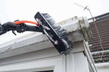 Kirli, tıkanmış, beyaz plastik pvc olukları temizleyen ve yosunlu yeşil plastik fasialarla boruları boşaltan bir fırça. Tıkanmış kanallar ve oluklar için pencere temizleyicileri ve iyi drenaj için düzenli bahçe bakımı gerekiyor.