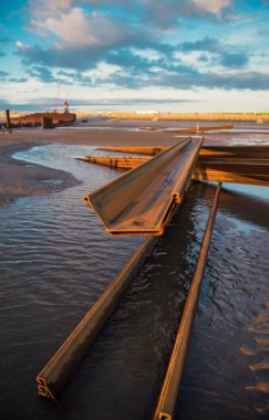 Büyük paslı çelik inşaat sütunları güneşli bir kumsalda, mavi gökyüzü ve sakin dalgalarla. Çelik kirişler paslanmış. Sahilde inşaat ve inşaat. Geliştirme ve inşa