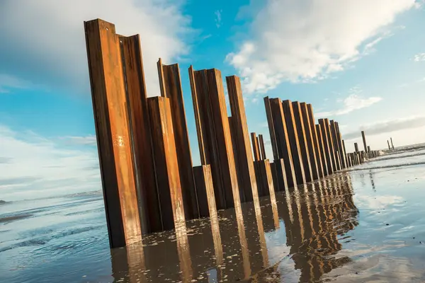 stock image Large rusty steel construction pillars in the sand on a sunny beach, with blue sky and calm waves. The steel girders are rusty. Building and construction on the coast. Development and building