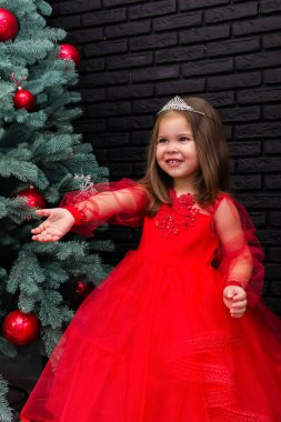 Little girl in red dress smiling by Christmas tree. Little beautiful girl in a red evening dress the Christmas tree. Girl in red in Christmas decorations. Christmas concept. New Year. Baby near a