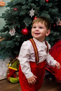 Kırmızı elbiseli küçük bir çocuk stüdyodaki şöminenin yanındaki yapay karda eğleniyor. Kırmızı Noel kostümü giymiş küçük bir çocuk süslü bir Noel ağacının yanında eğleniyor. Bebek oyuncağı