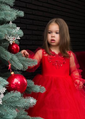 Little girl in red dress smiling by Christmas tree. Little beautiful girl in a red evening dress the Christmas tree. Girl in red in Christmas decorations. Christmas concept. New Year. Baby near a