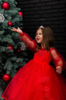 Little girl in red dress smiling by Christmas tree. Little beautiful girl in a red evening dress the Christmas tree. Girl in red in Christmas decorations. Christmas concept. New Year. Baby near a