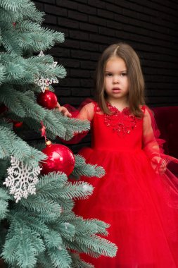 Little girl in red dress smiling by Christmas tree. Little beautiful girl in a red evening dress the Christmas tree. Girl in red in Christmas decorations. Christmas concept. New Year. Baby near a