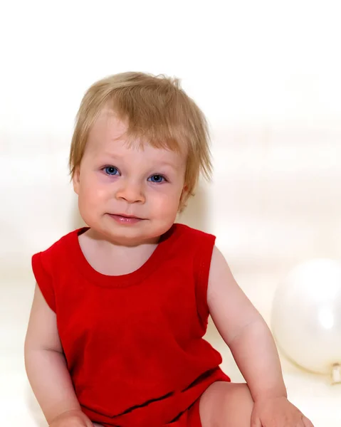 One-year-old toddler with blonde hair in red bodysuit sitting and smiling on white background with inflatable balloons. Wonderful kid sat down to rest. Baby posing isolated over light background.