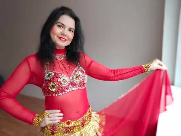 A dancer in a red suit is dancing an oriental dance on a white background. A professional dancer dances oriental belly dance. Sexy woman in red dress dances a seductive dance in a white studio