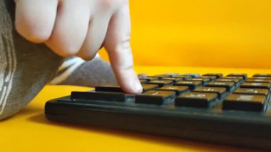 Küçük bir çocuk sarı arka planda bir hesap makinesine güveniyor. Çocuk parmaklarını tuşlara basar ve evde matematik çalışmayı hesaplar. Çocuk hesap makinesiyle aritmetik hesaplamalar yapıyor. Yüksek kalite 4K