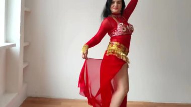 Kırmızı elbiseli bir dansçı beyaz arka planda doğulu bir dans ediyor. Profesyonel bir dansçı oryantal göbek dansı yapar. Kırmızı elbiseli seksi kadın beyaz bir stüdyoda baştan çıkarıcı bir dans yapıyor.