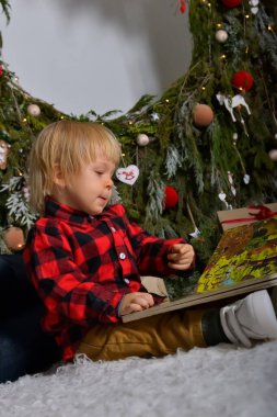 Gülümseyen küçük çocuk Noel ağacının yanında kitap okuyor. Evde Noel ağacının yanında oturan bir çocuk. Beyler. Aile Noel Kavramı. 