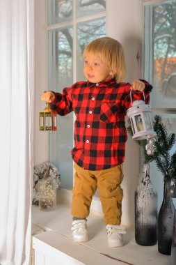 Çocuk elinde bir noel cücesiyle pencere eşiğinde oturuyor ve elinde bir Noel feneri ve yılbaşı hediyesi tutuyor, elinde bir kutu var. Aile Noel Kavramı. 