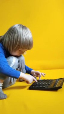 Küçük bir çocuk sarı arka planda bir hesap makinesine güveniyor. Çocuk parmaklarını tuşlara basar ve evde matematik çalışmayı hesaplar. Çocuk hesap makinesiyle aritmetik hesaplamalar yapıyor. Yüksek kalite FullHD
