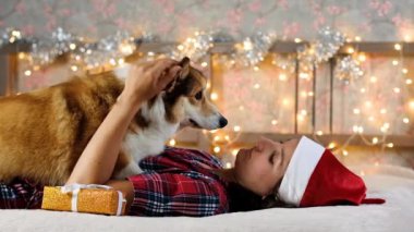 Woman in Santa hat plays with happy Corgi dog. Gifts and Christmas decorations against the garland lights background. New Year and Christmas concept. Waiting for the holiday