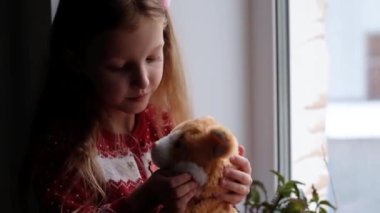 Küçük kız oturuyor, oyuncağına sarılıyor ve pencereden bakıyor. Kış zamanı ve evde kalma konsepti