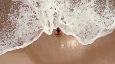 Mavi deniz dalgalarının yanındaki beyaz kumda bikinili genç bir kadın. Havadan çekim. Çarpıcı tropik plaj manzarası. Kız bir dronu kontrol ediyor ve kendi fotoğraflarını çekiyor..